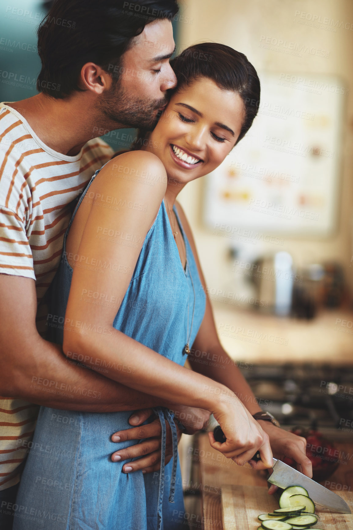 Buy stock photo Man, woman and kiss with cooking in kitchen, smile and care with diet, hug and prepare lunch in home. Indian people, happy couple and embrace with cucumber, love and meal prep for dinner in house