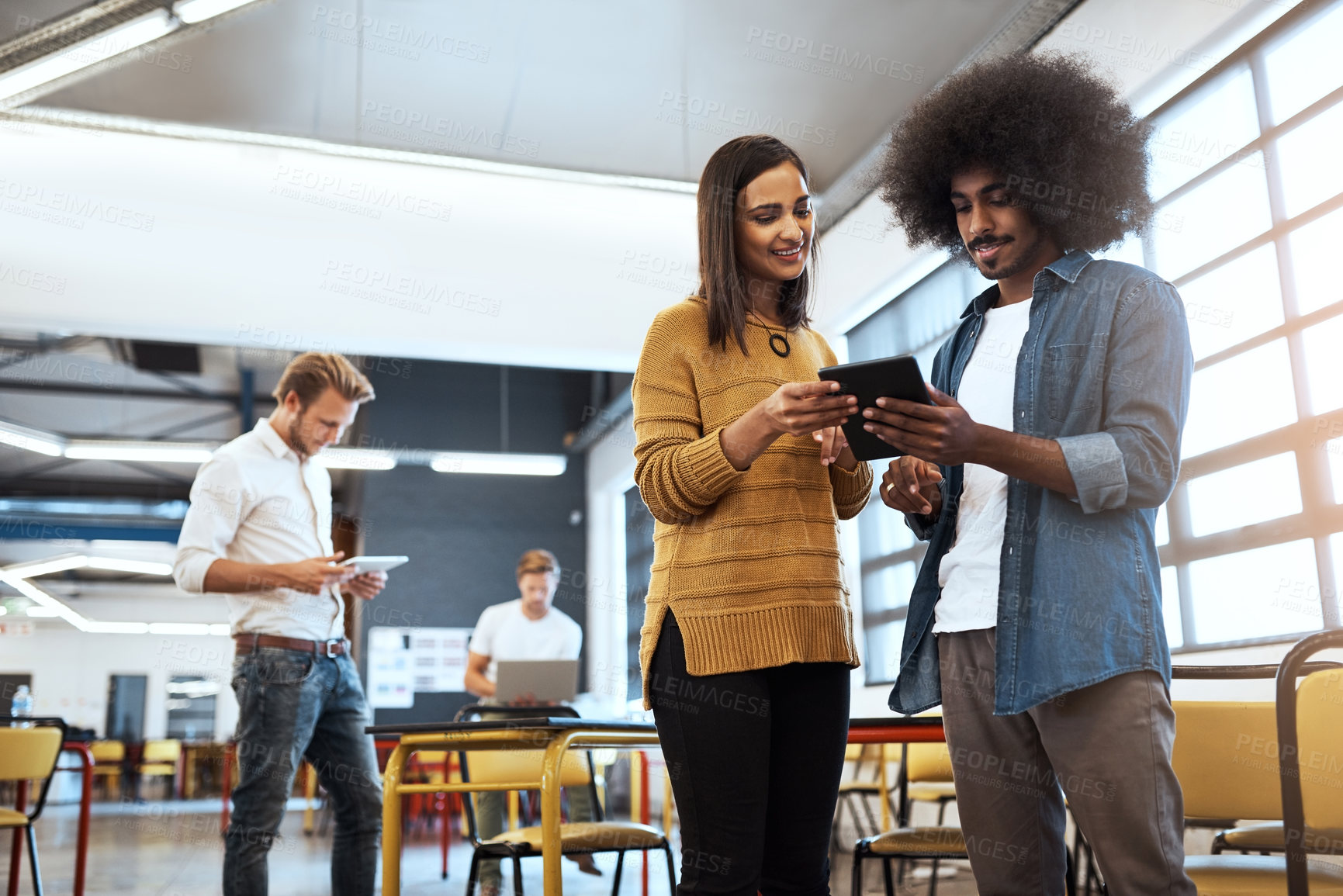 Buy stock photo Cropped shot of two young businesspeople looking at a tablet in the office with their colleagues in the background