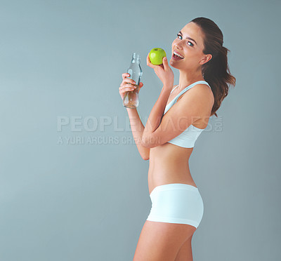 Buy stock photo Studio shot of a healthy young woman holding a glass water bottle and an apple against a grey background