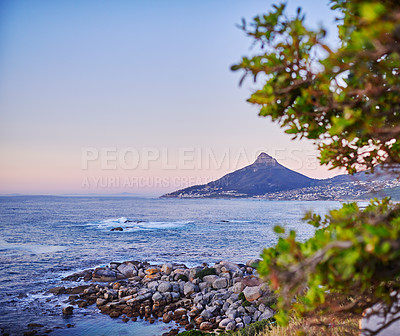 Buy stock photo Ocean view on a shallow rocky shoreline with Lion's Head, Table Mountain National Park in Cape Town, South Africa in the background. Quiet calm beach during sunset on a beautiful summer evening