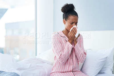 Buy stock photo Shot of a sickly young woman blowing her nose with a tissue in her bedroom at home