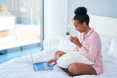 Buy stock photo Shot of an attractive young woman drinking coffee while using her laptop in bed at home