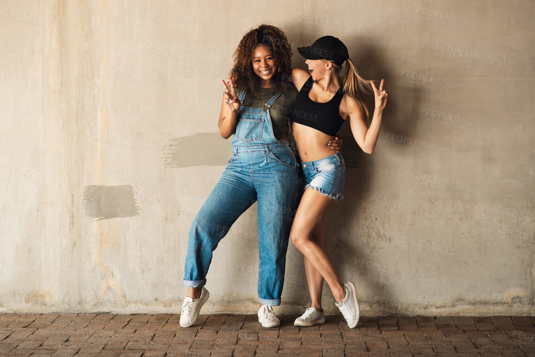 Buy stock photo Portrait of two cheerful young women posing for for a photo while leaning against a wall outside during the day