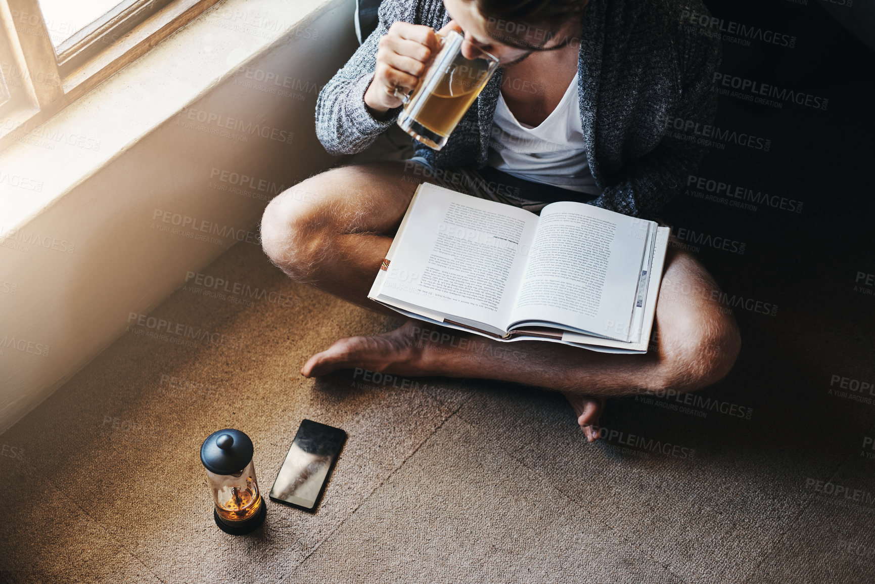 Buy stock photo High angle shot of a young man drinking tea and reading a book while sitting on the floor at home