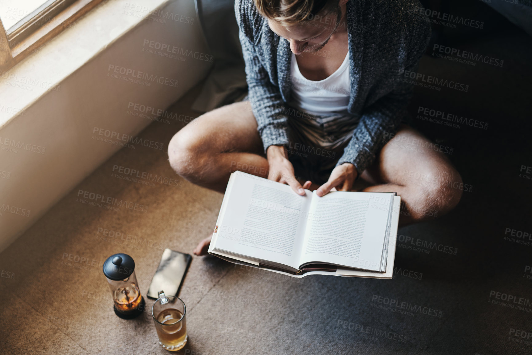 Buy stock photo High angle shot of a young man reading a book while sitting on the floor at home
