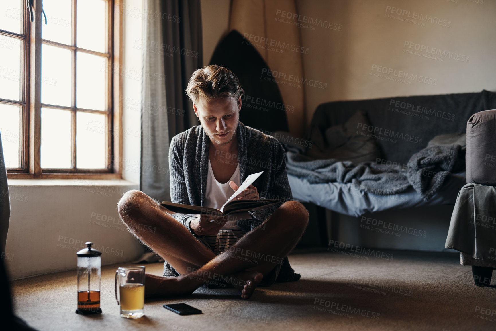 Buy stock photo Shot of a young man reading a book while sitting on the floor at home