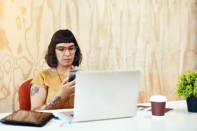 Buy stock photo Shot of a young designer using her cellphone while working on a laptop in an office