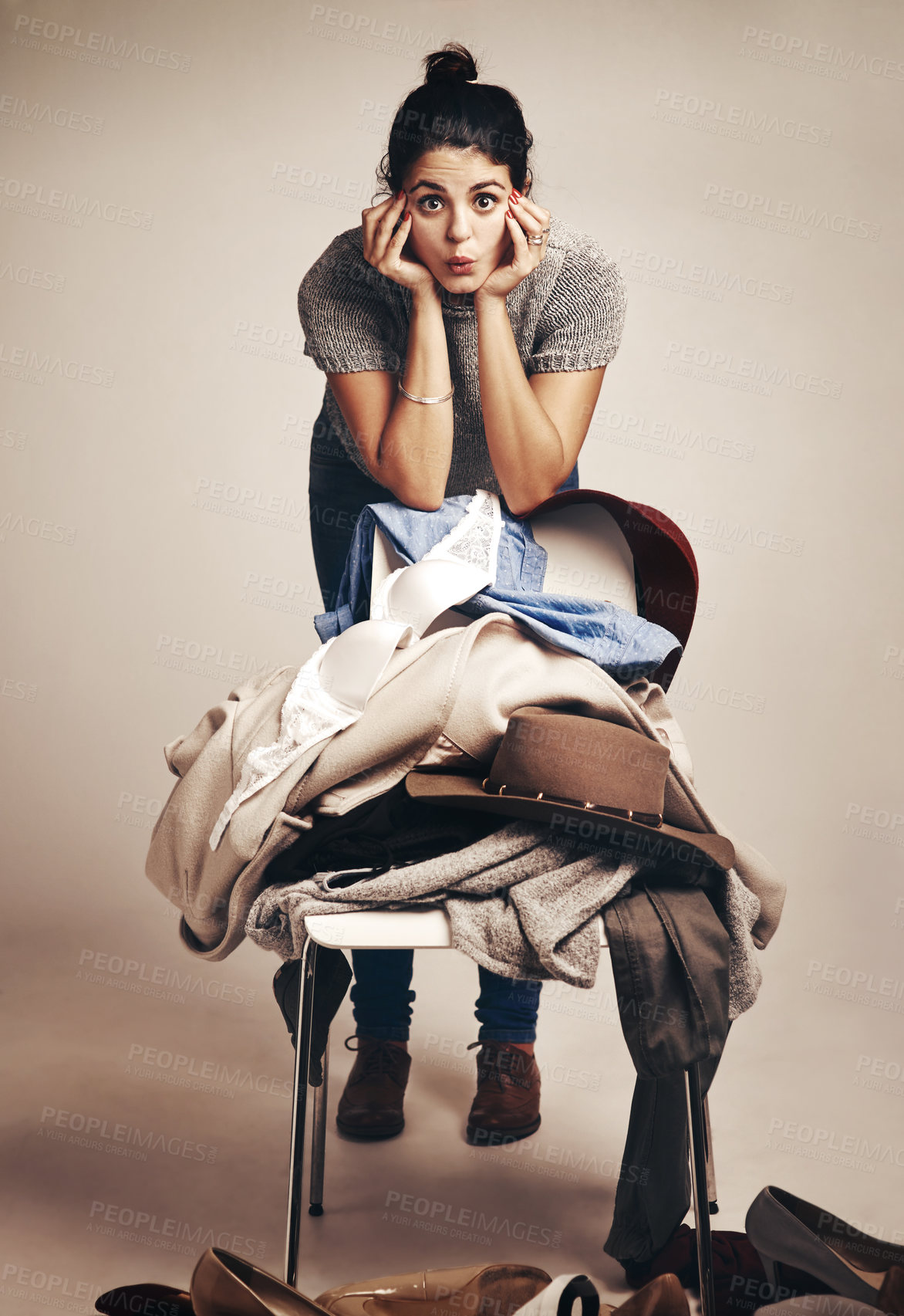 Buy stock photo Studio shot of a young woman choosing clothing piled on a chair against a brown background
