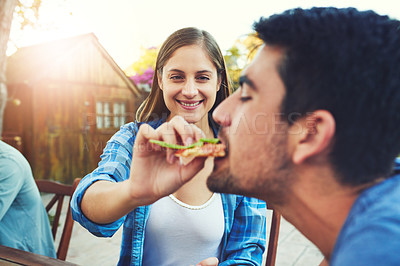Buy stock photo Shot a cheerful young man being fed pizza by his girlfriend in an outdoor gathering amongst friends
