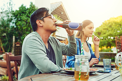 Buy stock photo Shot of a young man having a sip of a beer while sitting around a table in an outdoor gathering amongst friends
