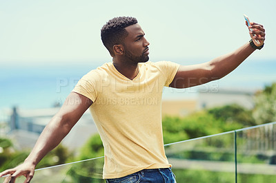 Buy stock photo Shot of a handsome young man taking a selfie with his cellphone while relaxing outdoors on holiday