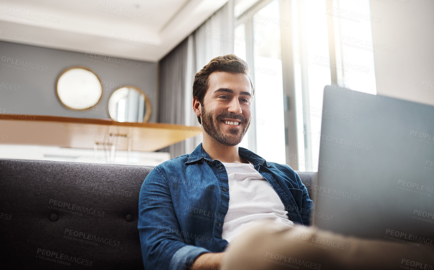 Buy stock photo Shot of a handsome young man using his laptop while relaxing on a sofa at home