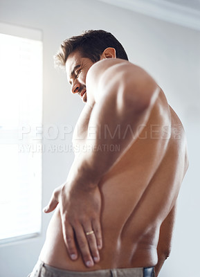Buy stock photo Rearview shot of a shirtless young man holding his back in pain while relaxing at home