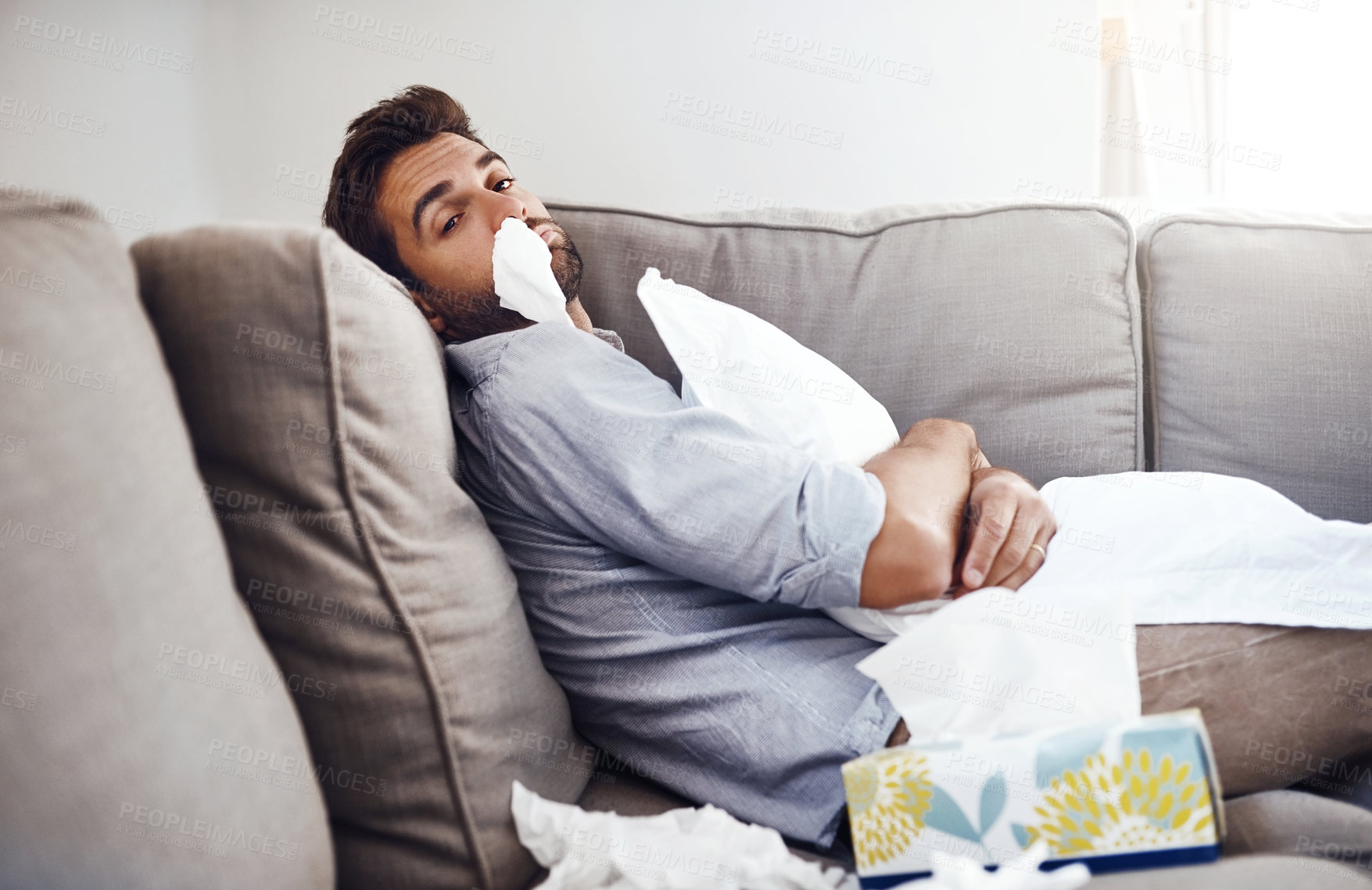 Buy stock photo Portrait of a sickly young man with a tissue up his nose siting on a couch and holding a pillow at home