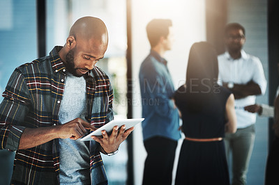 Buy stock photo Shot of a young businessman using his digital tablet in a modern office with his colleagues in the background