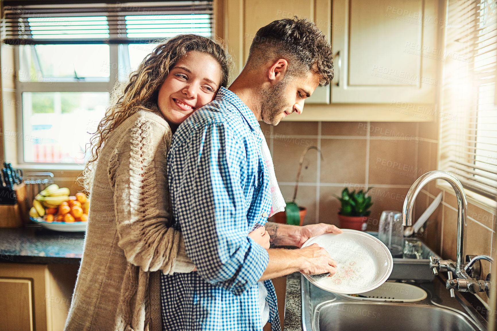 Buy stock photo Shot of a young woman embracing her boyfriend from behind while he does the dishes
