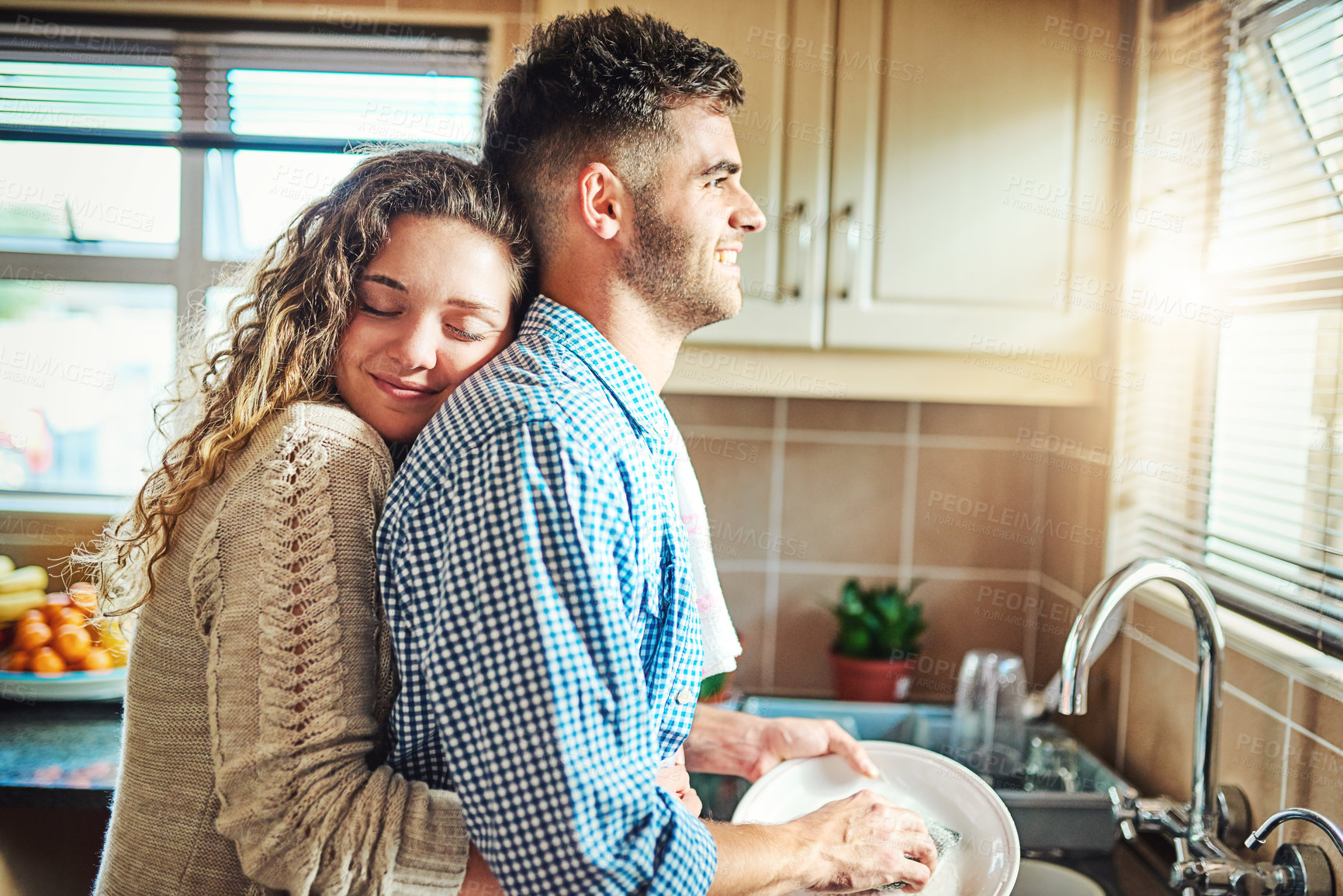 Buy stock photo Shot of a young woman embracing her boyfriend from behind while he does the dishes