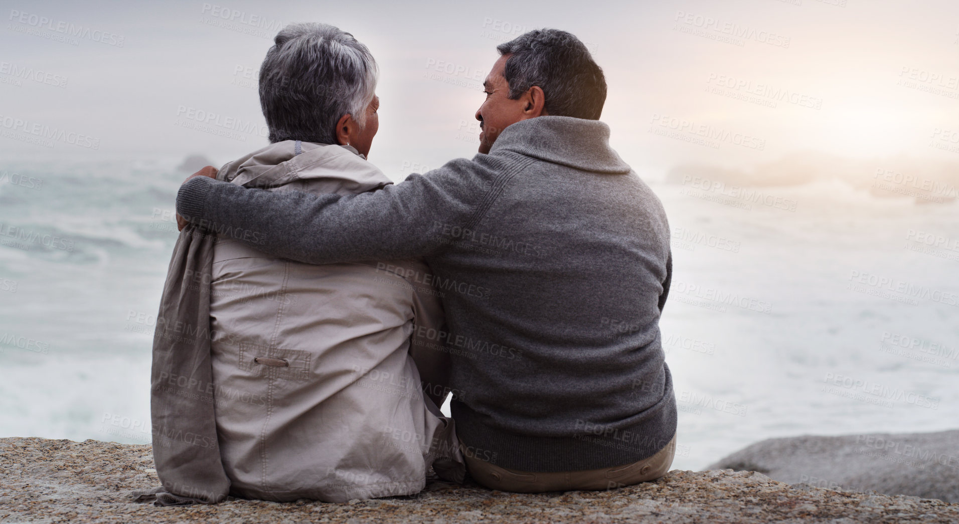 Buy stock photo Senior couple, hug and back at beach with sunset, waves and relax on rock with chat, love and bonding. Elderly woman, old man and talking with embrace by ocean for connection, vacation and retirement