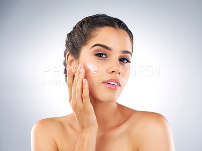 Buy stock photo Studio shot of a beautiful young woman applying moisturizer to her face against a grey background