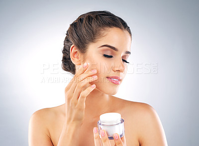 Buy stock photo Shot of a beautiful young woman holding a tub of moisturizer and applying it to her face in studio