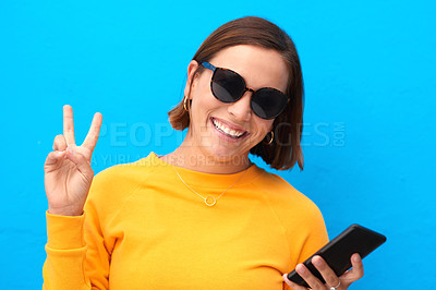 Buy stock photo Girl, peace sign and phone with sunglasses in studio for social media, casual fashion and trendy aesthetic. Smile, woman and v gesture of shades, mobile networking and cool style on blue background