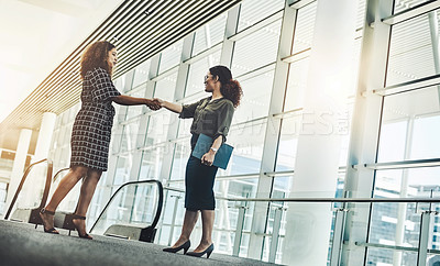 Buy stock photo Low angle shot of two attractive young businesswomen shaking hands while standing in a modern workplace