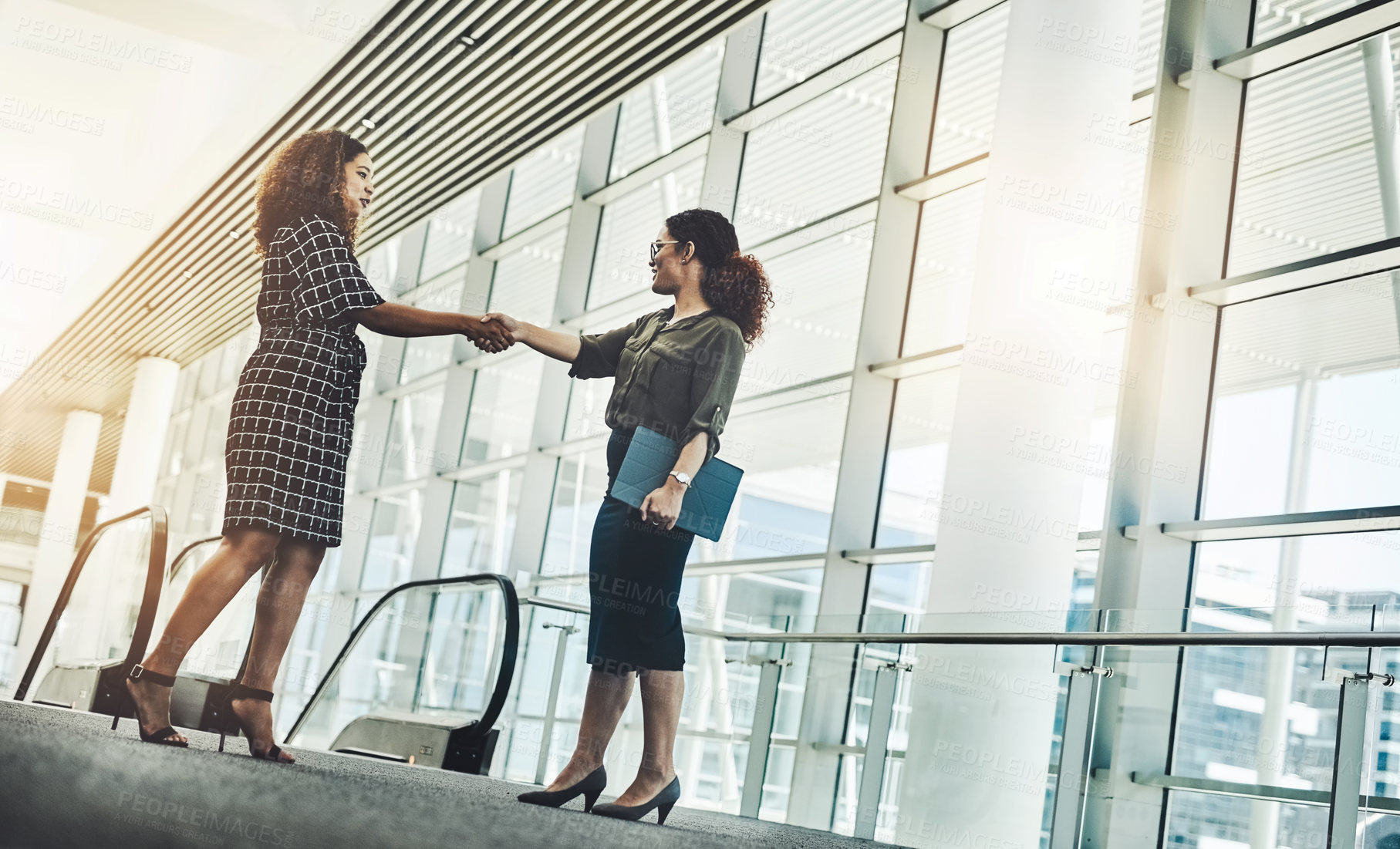 Buy stock photo Low angle shot of two attractive young businesswomen shaking hands while standing in a modern workplace