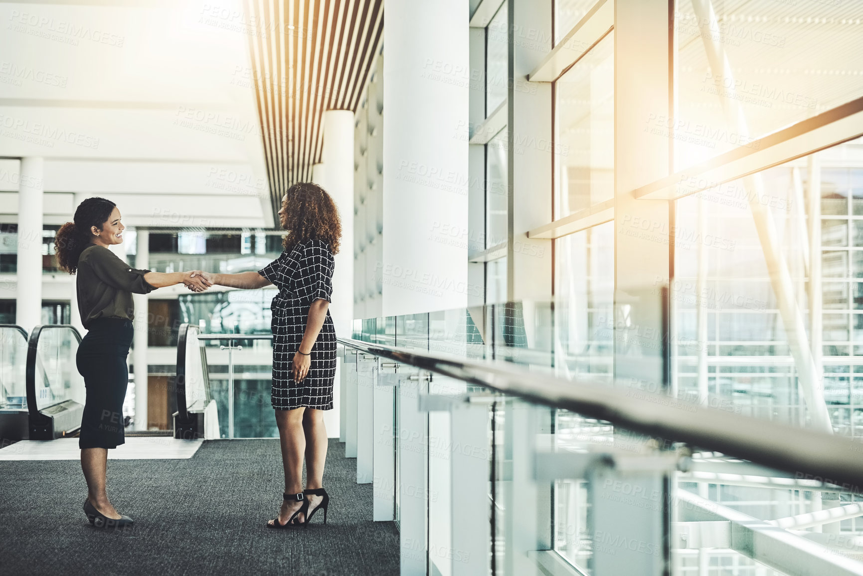 Buy stock photo Full length shot of two attractive  young businesswomen shaking hands while standing in a modern workplace