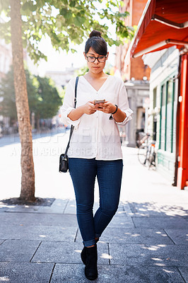 Buy stock photo Shot of a beautiful young woman walking on a sidewalk past shops while texting on her cellphone outside in a town during the day