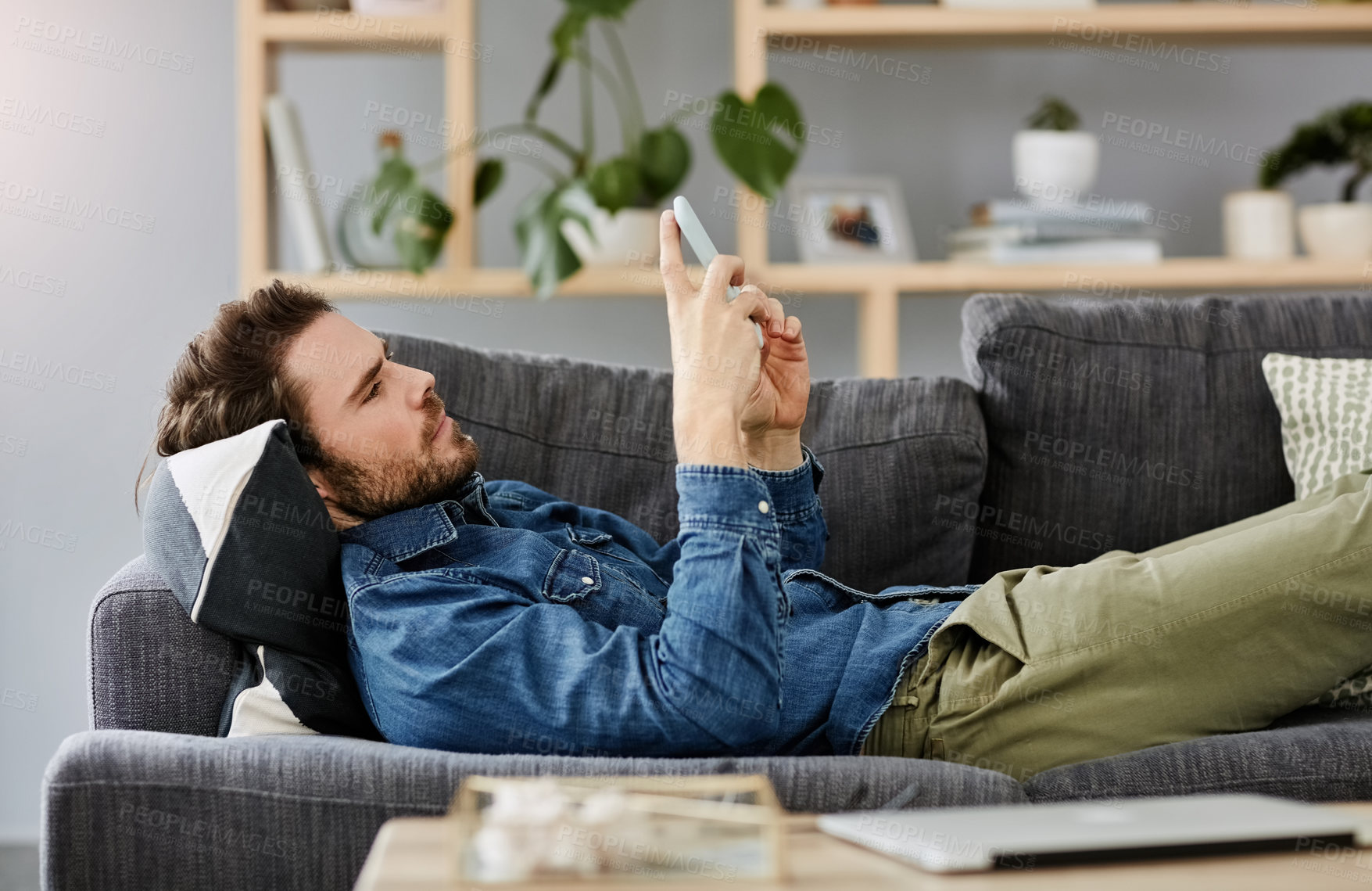 Buy stock photo Cropped shot of a handsome young man using a smartphone while lying on a couch at home