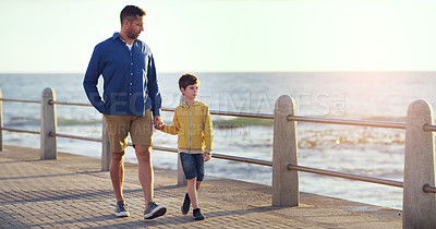 Buy stock photo Shot of a father and son holding hands while walking on a walkway near the beach