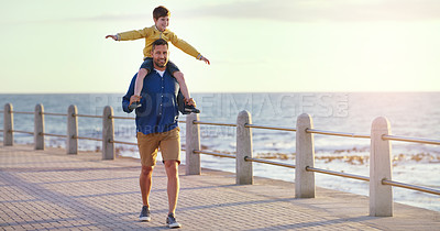 Buy stock photo Shot of a cheerful father carrying his son on his shoulders while taking a walk on walkway near the beach
