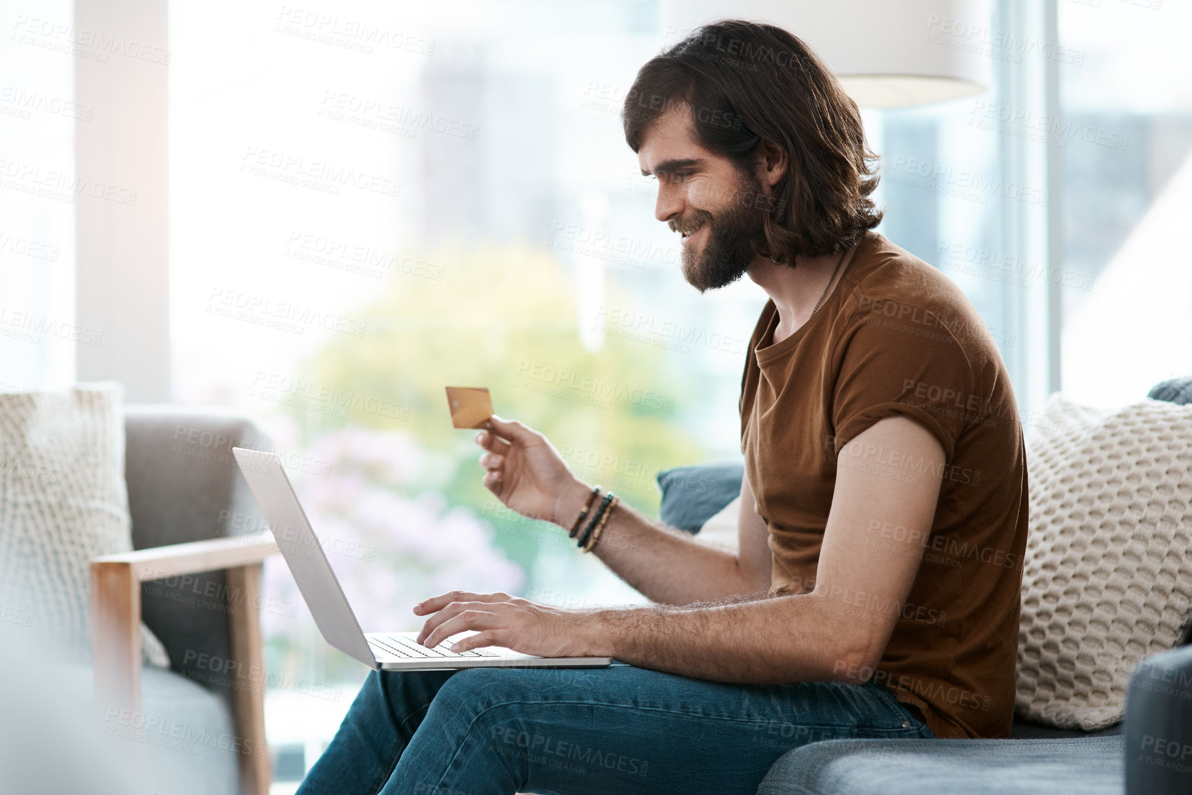 Buy stock photo Shot of a handsome young man holding a credit card and using his laptop while relaxing in his living room