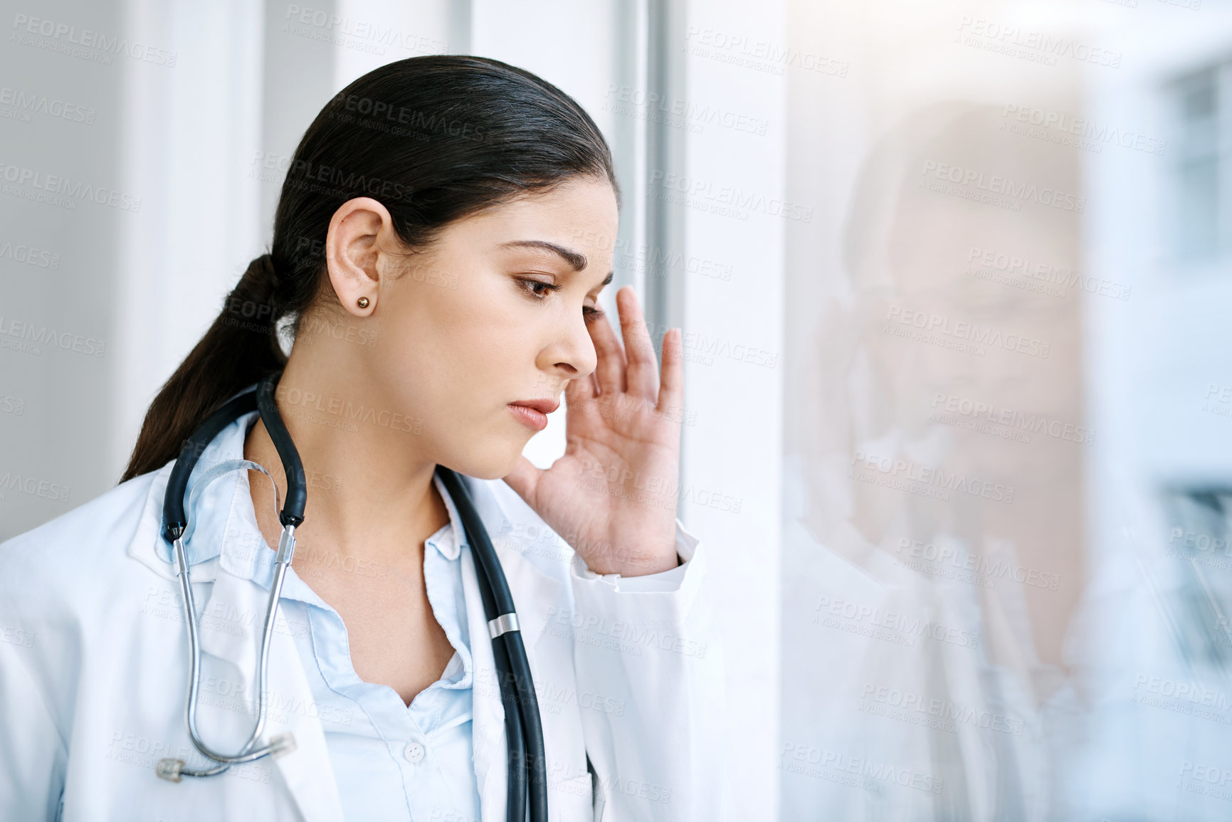 Buy stock photo Shot of a young female doctor looking stressed out while standing at a window in a hospital