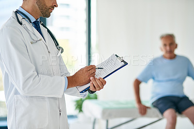 Buy stock photo Cropped shot of an unrecognizable young doctor filling out a file with his elderly patient in the background