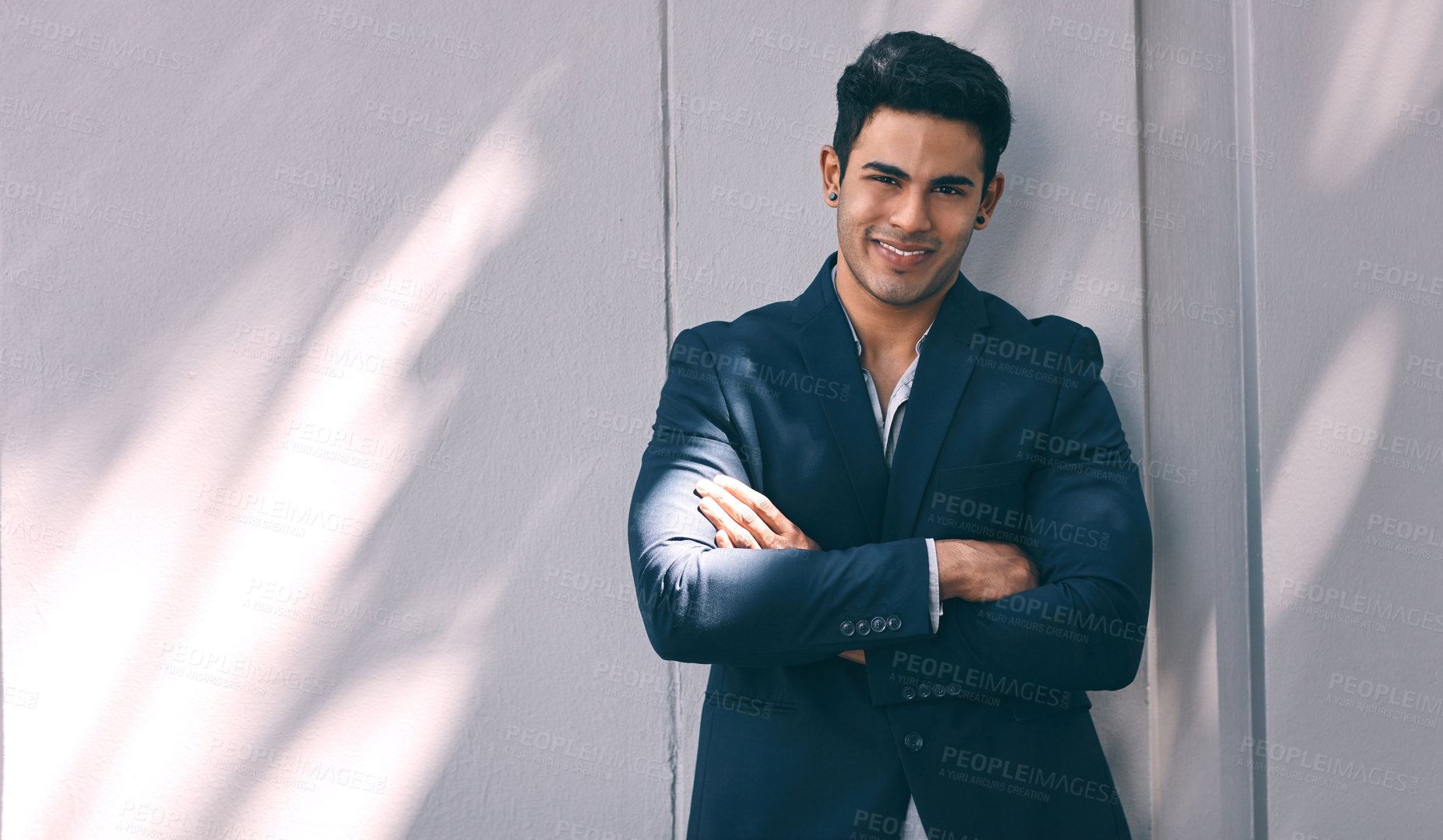 Buy stock photo Portrait of a handsome young businessman standing against a grey wall with his arms folded