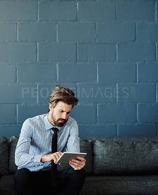 Buy stock photo Cropped shot of a handsome young businessman using his tablet while sitting on a sofa in the office