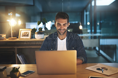 Buy stock photo Shot of a handsome young businessman smiling while working late at night on his computer in a modern office
