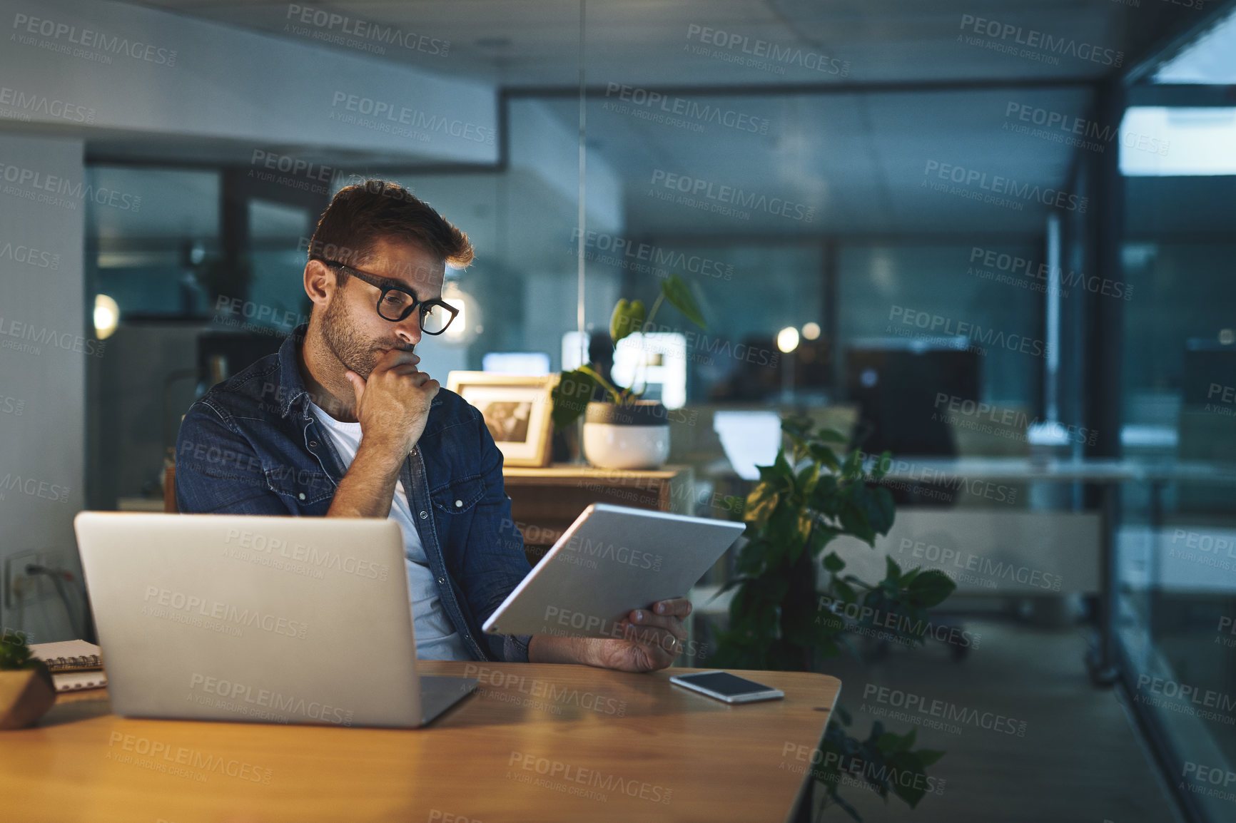 Buy stock photo Shot of a handsome young businessman working late at night on his tablet in a modern office