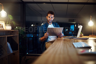 Buy stock photo Shot of a handsome young businessman smiling while working late at night on his tablet in a modern office