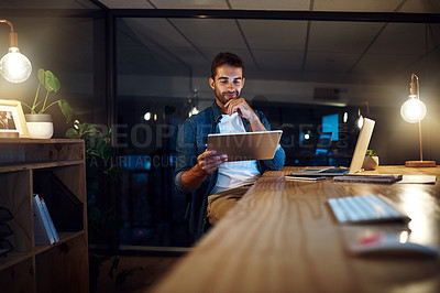Buy stock photo Shot of a handsome young businessman smiling while working late at night on his tablet in a modern office