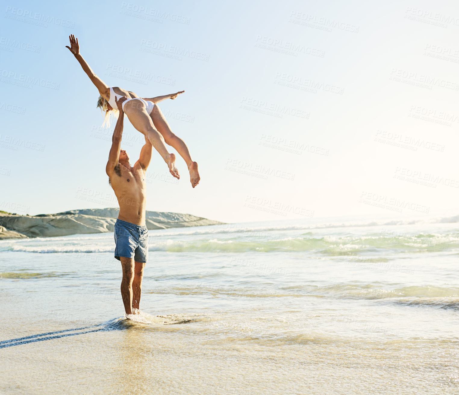Buy stock photo Shot of a young man lifting his girlfriend up into the air at the beach