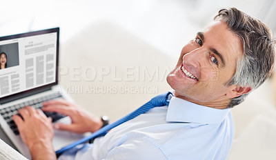 Buy stock photo Portrait of a mature businessman smiling and sitting on a sofa while working on his laptop indoors