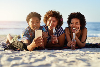Buy stock photo Shot of an attractive young trio of women laying down on the beach and taking selfies together during the day