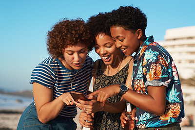 Buy stock photo Shot of three attractive young women huddled together and looking at a cellphone on the beach during the day