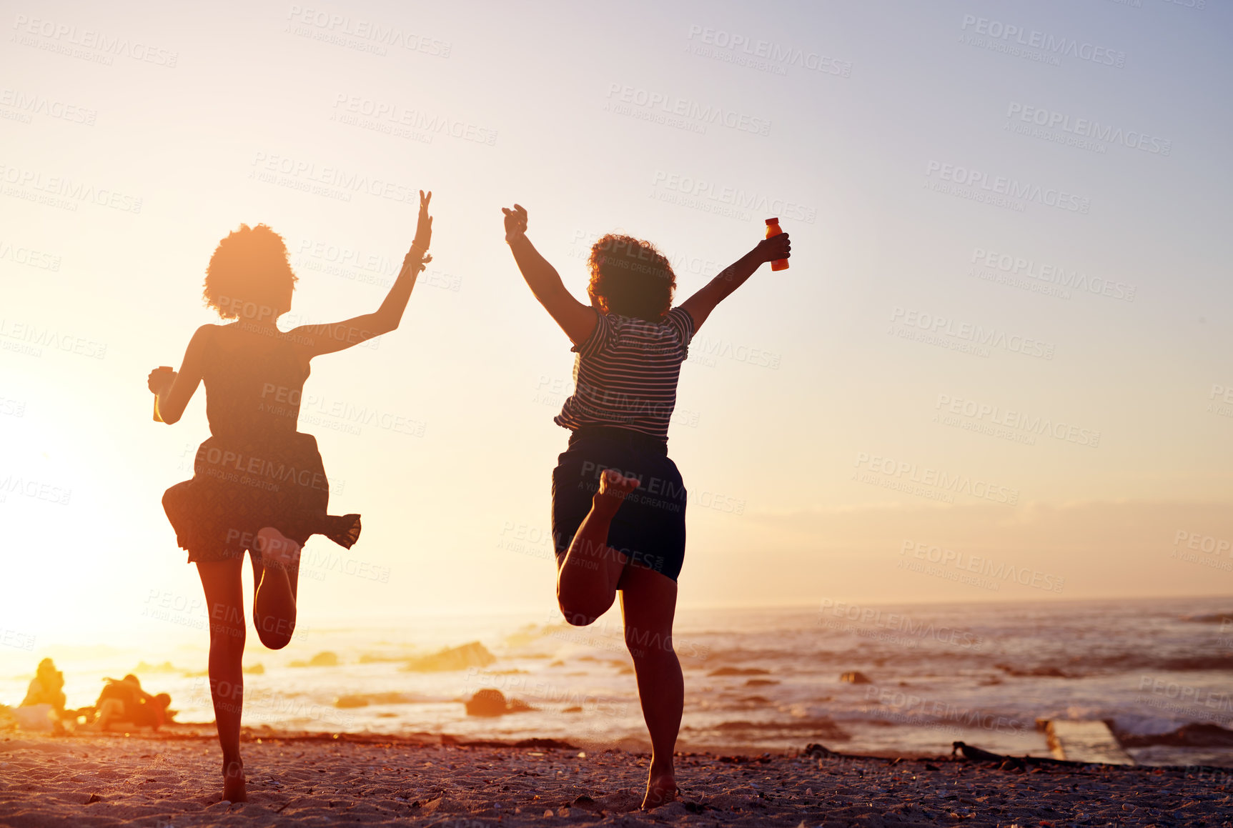 Buy stock photo Rearview shot of two unrecognizable young women jumping up in joy while walking along the beach at sunset