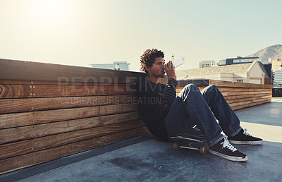 Buy stock photo Full length shot of a young man sitting down on his skateboard and drinking water at a skatepark