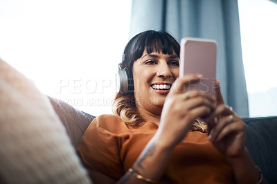 Buy stock photo Shot of a young woman wearing headphones while using her cellphone at home