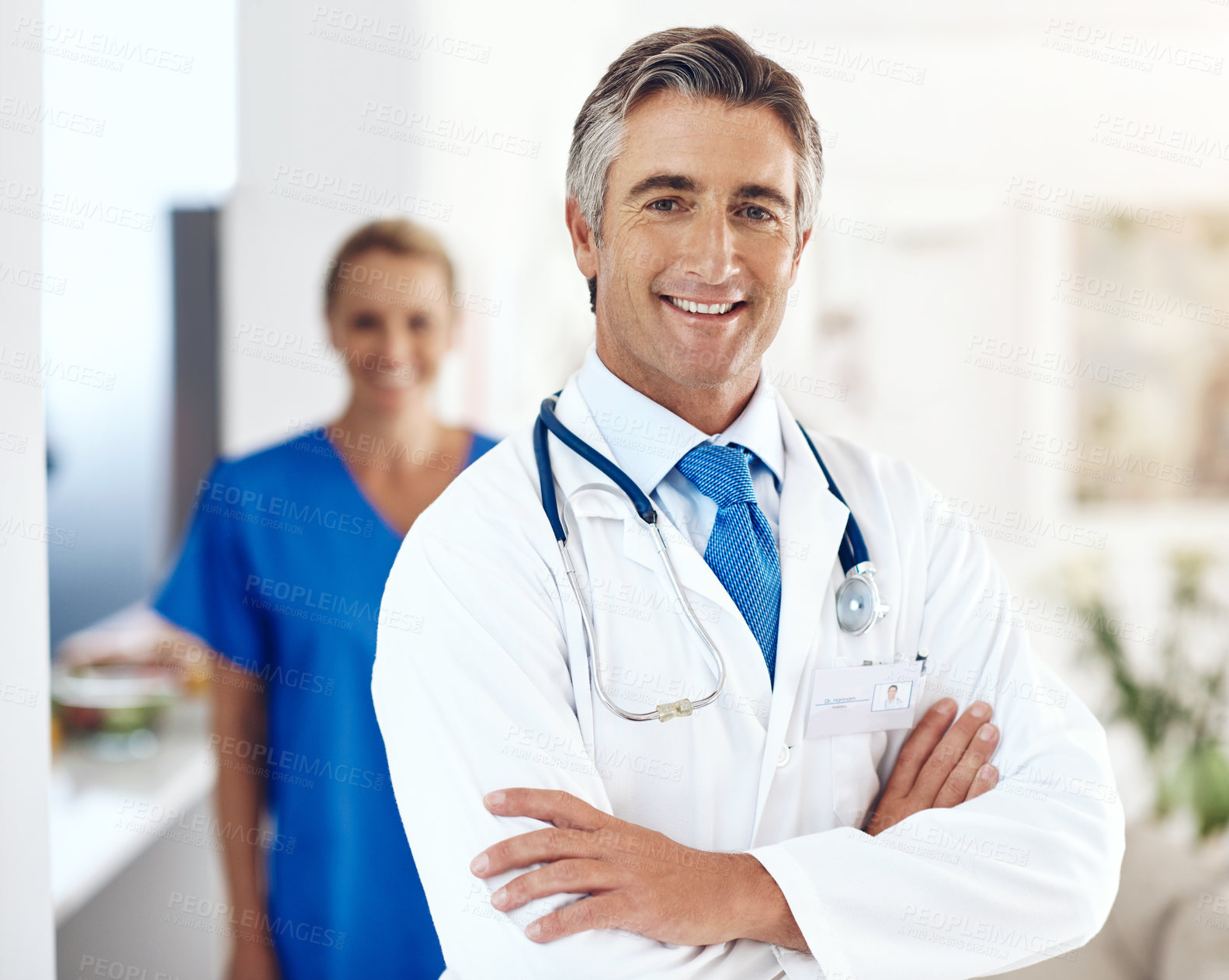 Buy stock photo Portrait of a male doctor standing with his arms crossed in the hospital with a female colleague in a the background