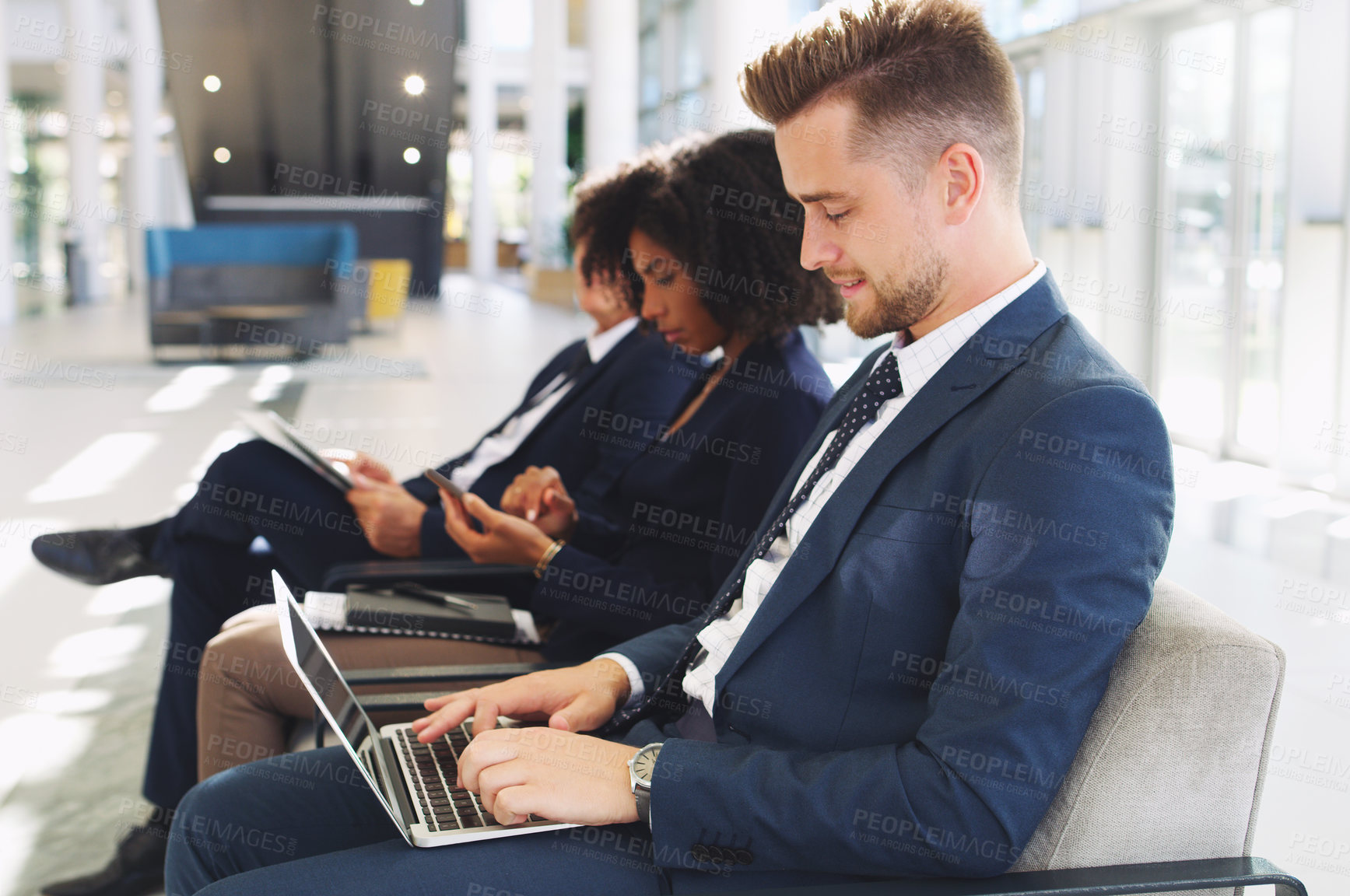 Buy stock photo Cropped shot of a handsome young businessman using a laptop while sitting in a line with his colleagues in the office